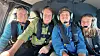 Fire personer med headset sidder tæt sammen i et fly eller helikopter og smiler.